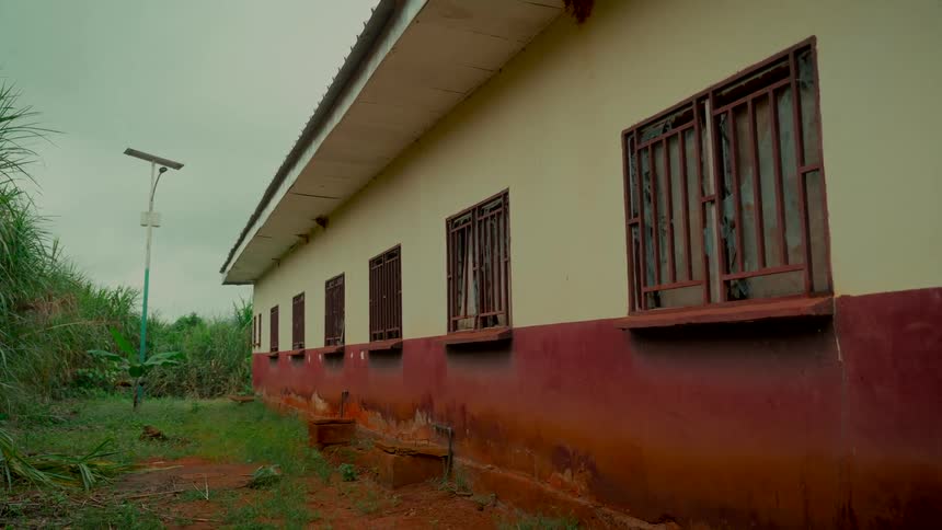 A rural building with barred windows and a solar-powered lamp stands amidst overgrown vegetation on an overcast day