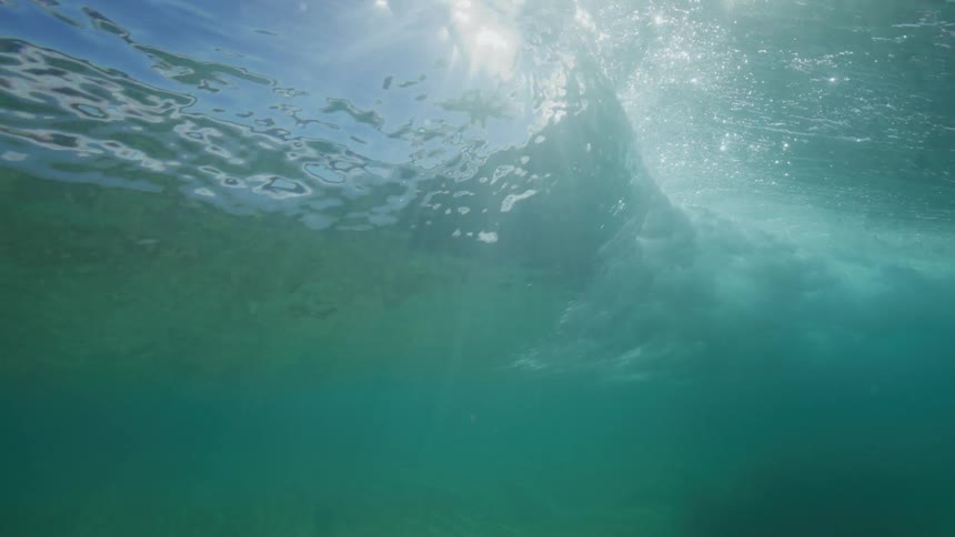 A slow-motion underwater view captures an ocean wave cresting and passing overhead as bright sunlight illuminates the turquoise water