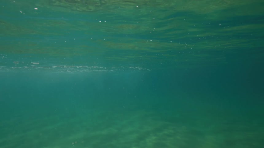 An underwater perspective of the tranquil ocean shows sunlight filtering through the calm, turquoise water to the sandy sea floor below
