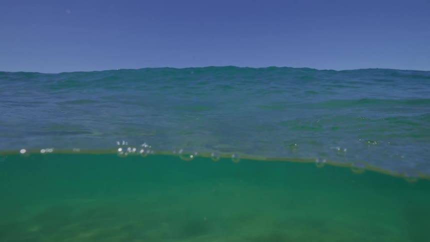 A serene, slow-motion over-under shot captures the gentle ripples on the ocean surface against a clear blue sky, with a view of the sandy seabed below