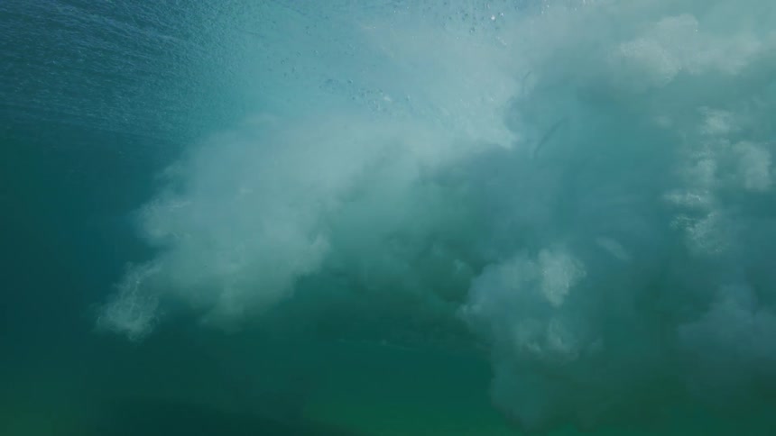 An underwater view captures a powerful ocean wave breaking in dramatic slow motion, revealing a swirling vortex of white foam and turquoise water