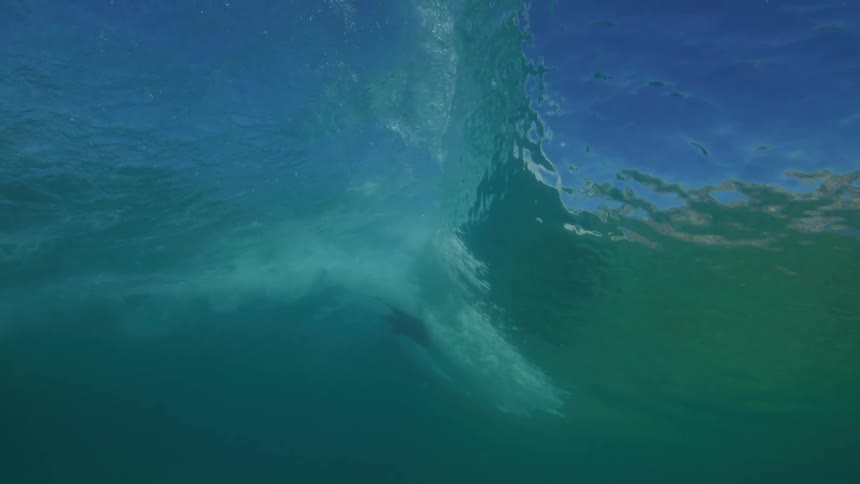 A slow-motion underwater view shows surfers paddling on the surface as a powerful ocean wave builds and breaks overhead