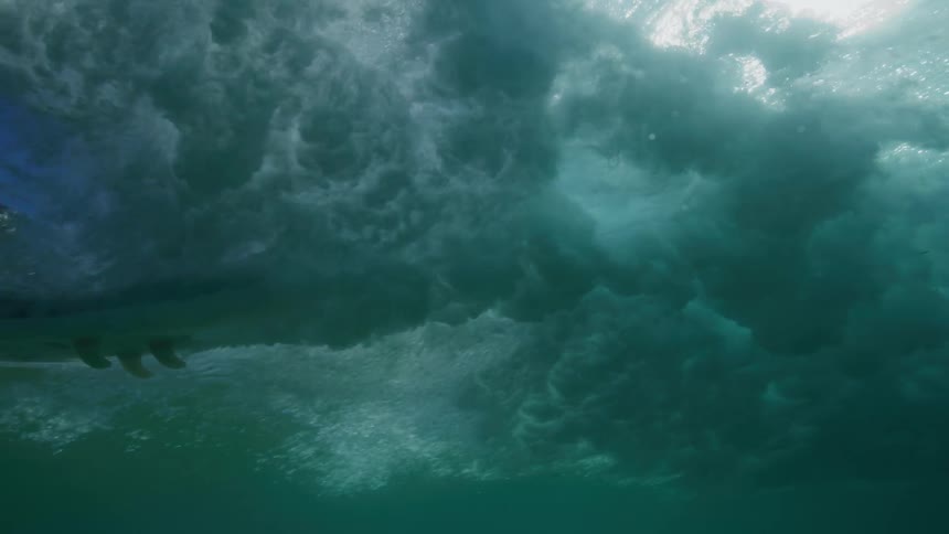 An immersive underwater view captures a surfer in slow motion carving through a crystal-clear wave, with brilliant sun rays illuminating the turbulent water from above