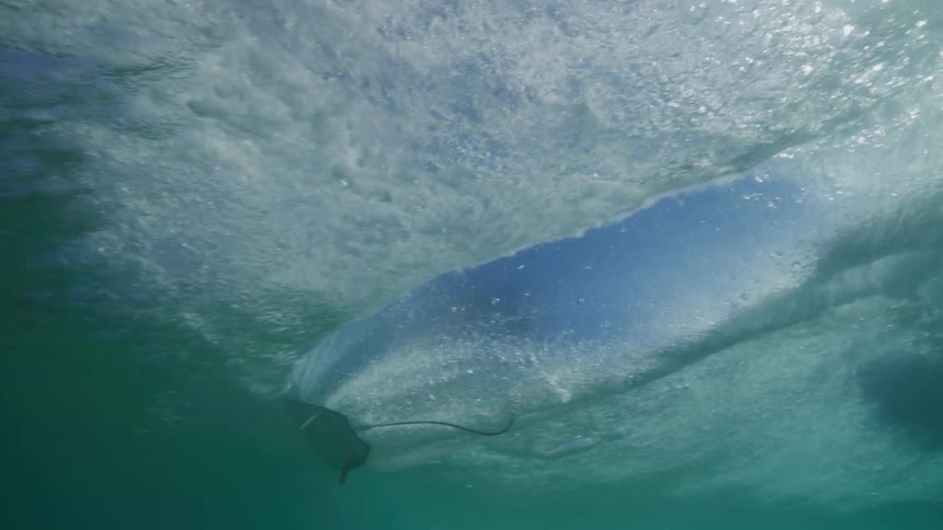 Slow motion underwater shot of a surfer paddling and riding a wave with bright sunlight filtering through the ocean