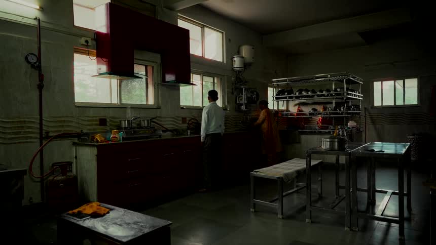 Wide, static shot of two people working in a large, atmospheric institutional kitchen