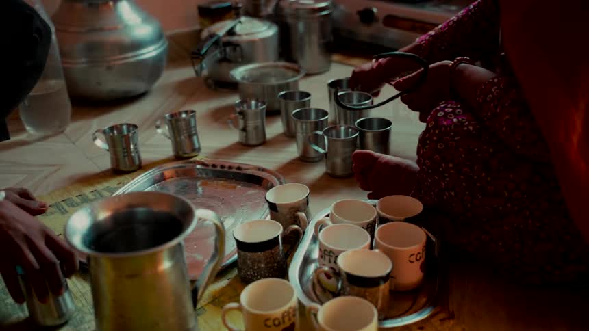 Women gather on the floor to prepare and serve hot drinks in a variety of mugs in a simple, rustic home