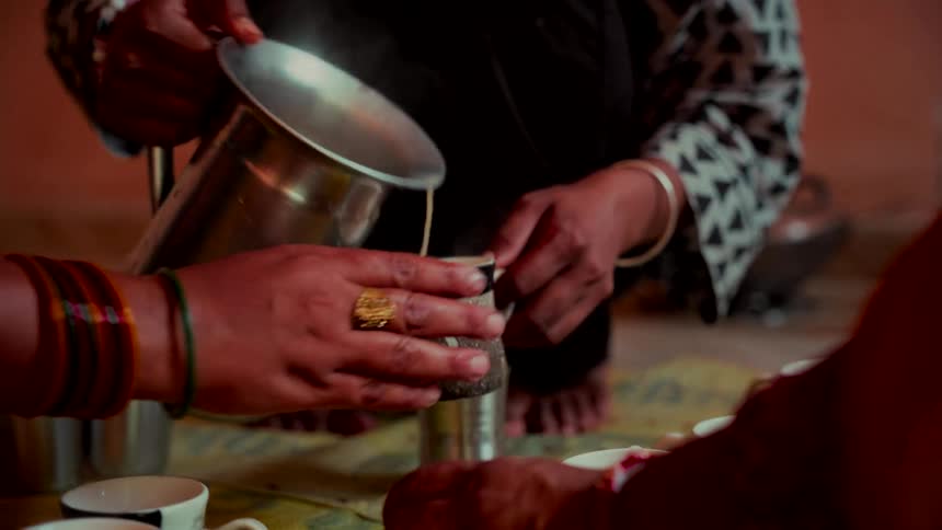 A woman pours hot steaming tea from a metal pitcher into a mug in slow motion during a traditional gathering