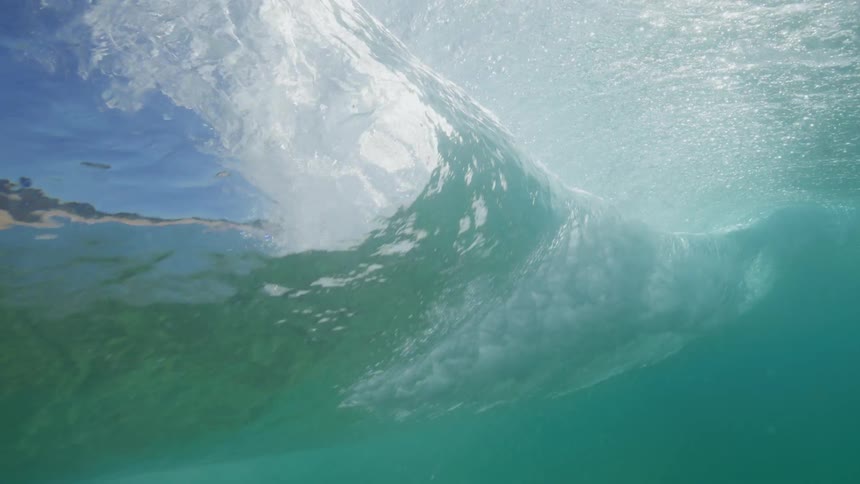 A stunning underwater slow-motion shot captures a turquoise ocean wave forming and cresting against the bright sun