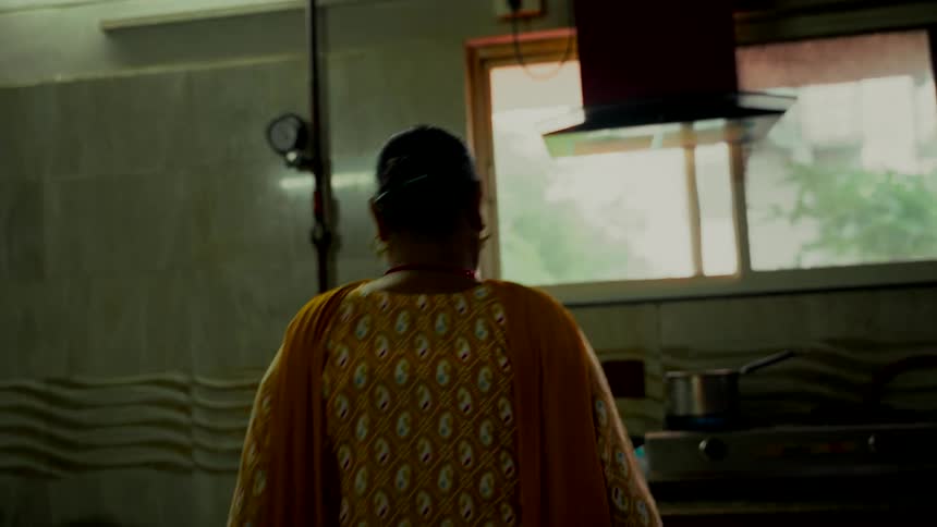 From behind, an Indian woman in a colorful traditional outfit stands at her stove preparing a meal in a modest kitchen
