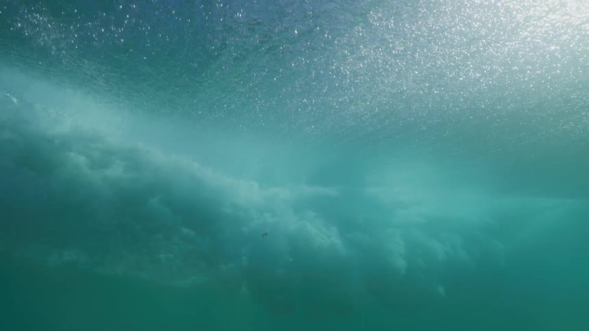 An immersive slow-motion underwater view of a wave cresting overhead, revealing the sunlit sky through the shimmering water's surface