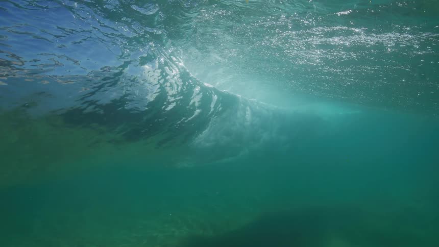 An underwater slow-motion view shows a turquoise ocean wave curling and breaking as sunlight streams through the surface