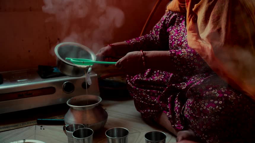 An Indian woman in traditional clothing sits on the floor and slowly strains steaming hot chai tea into a metal pot in slow motion