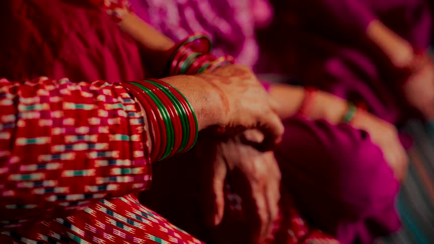 An elderly woman's hands, adorned with henna and vibrant bangles, rest patiently during a traditional ceremony