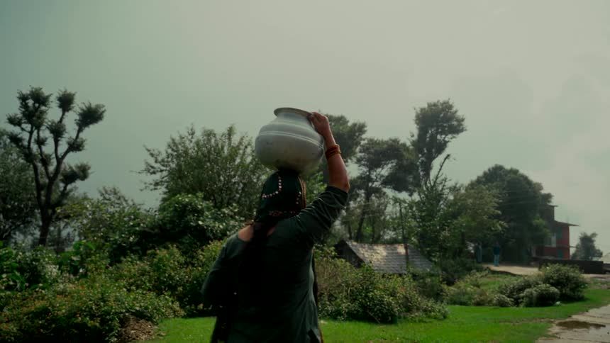 A woman seen from behind balances a large water pot on her head while walking through a lush, green rural village