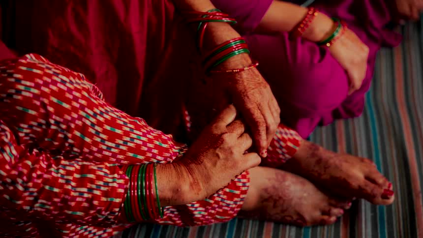 A detailed close-up of two women in colorful traditional clothing, their hands and feet adorned with intricate henna designs and vibrant bangles