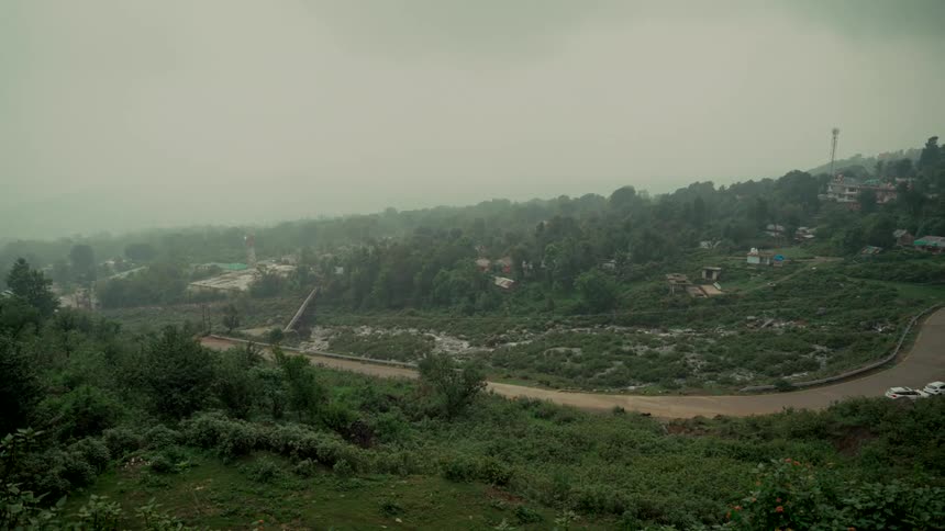 A wide shot captures a lush green valley with a winding road and a small village during a foggy, rainy day
