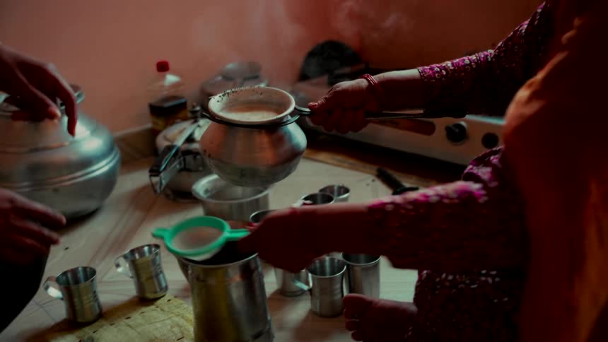 An Indian woman in traditional clothing prepares hot, steaming masala chai in a modest kitchen