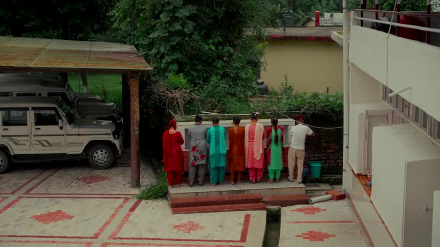 A high-angle view shows a group of people in vibrant, traditional Indian attire washing their hands at an outdoor station in a courtyard