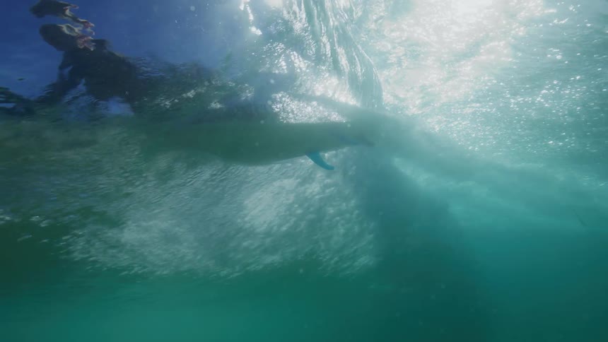 From beneath the surface, a surfer glides across a sunlit wave in a dramatic slow-motion underwater shot