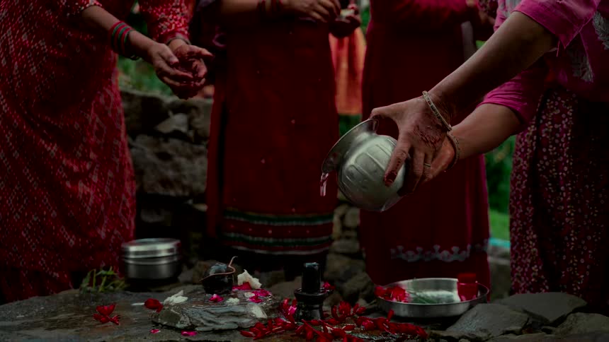A woman in traditional Indian clothing pours holy water from a silver pot over a Shiva Lingam during an outdoor Hindu puja ceremony
