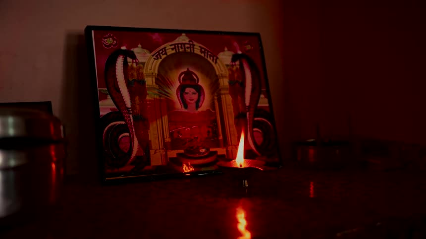 A traditional diya oil lamp flickers in the dark, illuminating a sacred picture of the Hindu serpent goddess, Nagni Mata