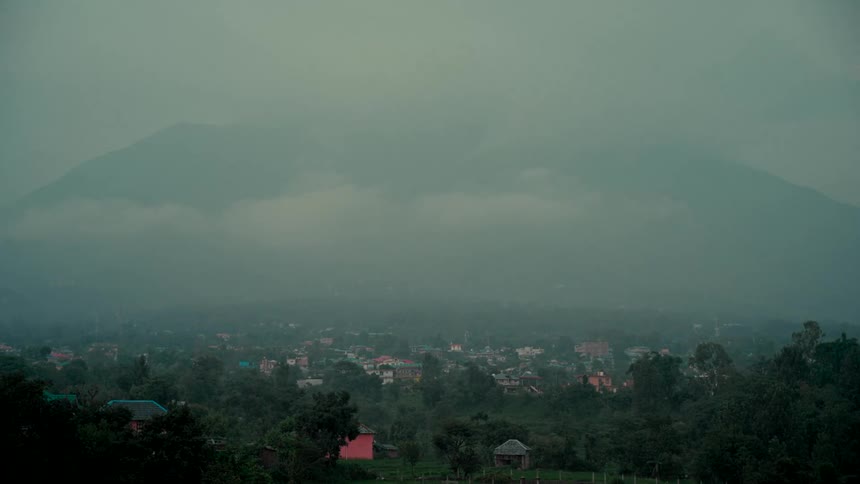 A moody and atmospheric wide shot captures a small town nestled in a lush valley under foggy mountains