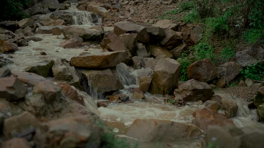 A small, muddy creek cascades over a bed of large rocks in a natural setting