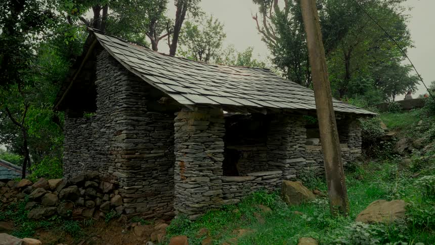 An old abandoned stone hut with a slate roof sits amidst lush green trees in a quiet, rural landscape