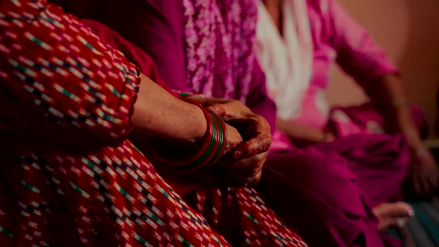 A close-up of an elderly woman's hands adorned with bangles, resting on her lap while dressed in colorful traditional clothing