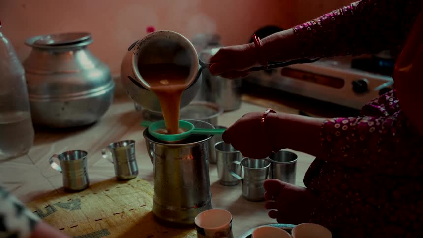 An Indian woman pours freshly brewed, steaming masala chai through a strainer into a metal pitcher in a rustic kitchen, preparing to serve