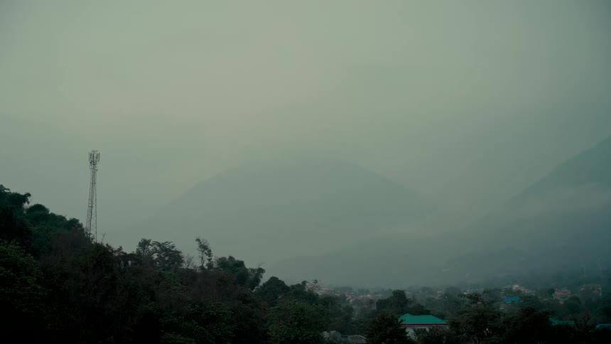 A foggy mountain range looms over a lush green valley with a telecommunications tower on an overcast day