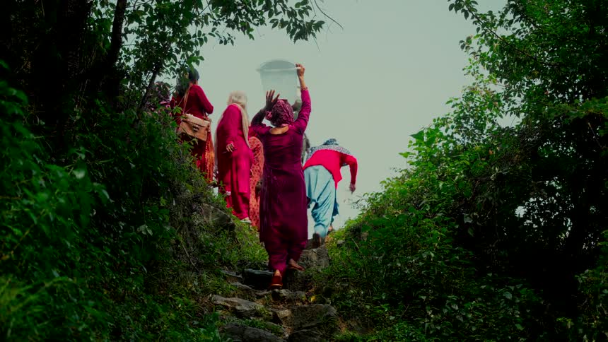 A group of women in vibrant traditional clothing carry water containers up a steep, verdant staircase in slow motion
