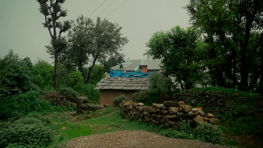 A traditional stone and mud house sits nestled in a lush green valley in the mountains on an overcast day