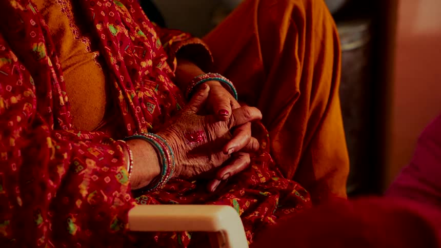 An elderly Indian woman with bangles on her wrists sits with her weathered hands clasped in her lap