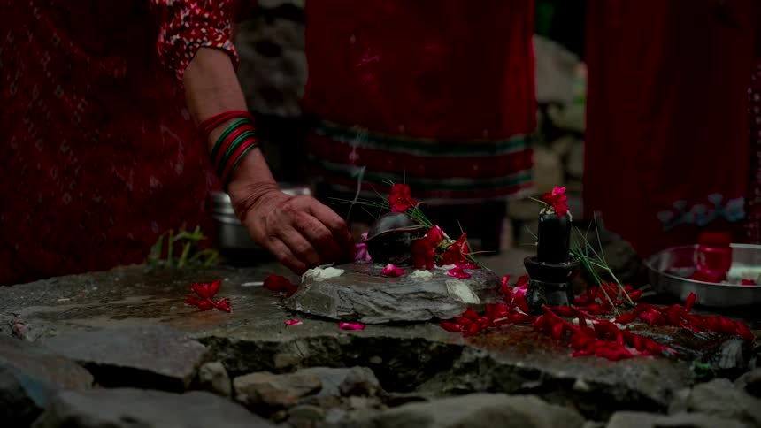 Women's hands perform a sacred Hindu Puja ritual, offering red flowers, rice, and incense to a Shiva Lingam on a stone altar