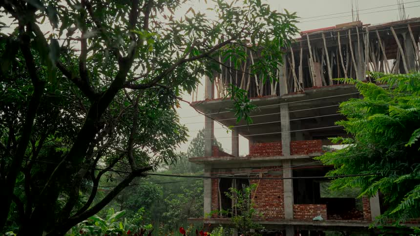An unfinished multi-story building is framed by lush green trees as gentle rain falls on the construction site