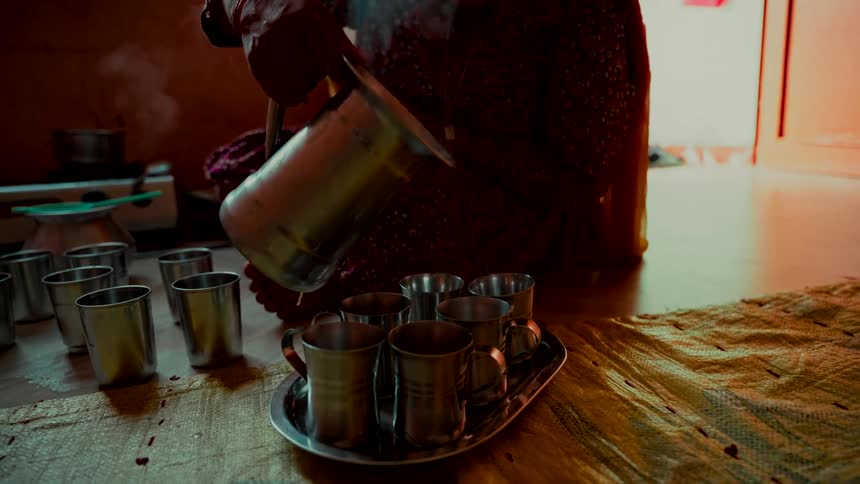 Slow motion shot of a woman pouring hot, steaming tea from a pitcher into traditional steel cups in a rustic home