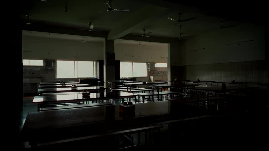 A static shot of a large, dimly lit, and empty institutional cafeteria with rows of tables and benches