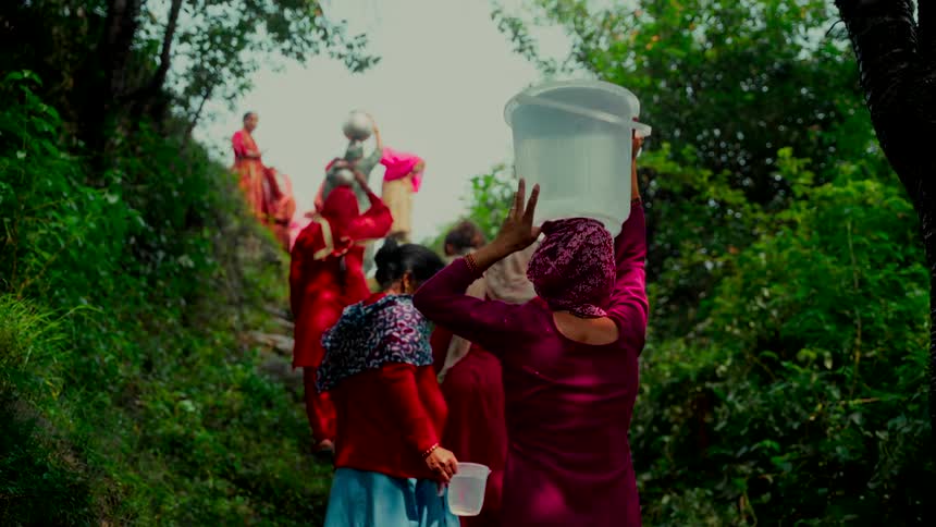 A group of women in traditional Indian attire carry buckets of water on their heads as they walk up a steep, rocky path through a lush green forest