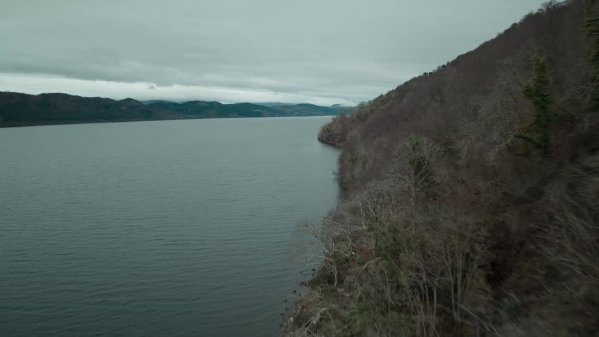 An aerial flight moves along the barren, forested coastline of a vast loch on a moody, overcast day