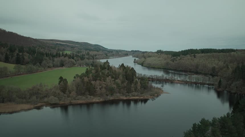 A drone gracefully ascends above a pine forest to reveal a serene, expansive lake and rolling hills under a cloudy sky