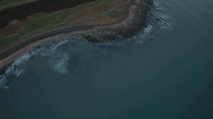 A static aerial top-down view of gentle ocean waves lapping against a rugged, rocky coastline on an overcast day