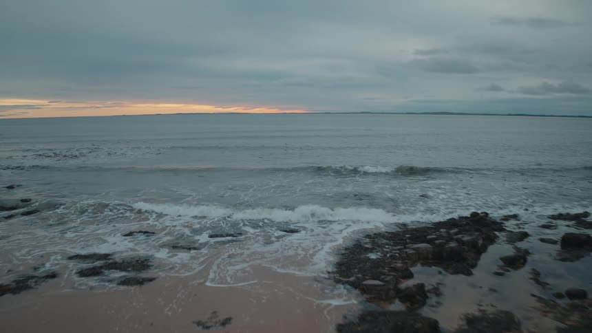 An aerial view glides over a rocky shoreline and out to the calm ocean as waves gently roll in under a cloudy twilight sky