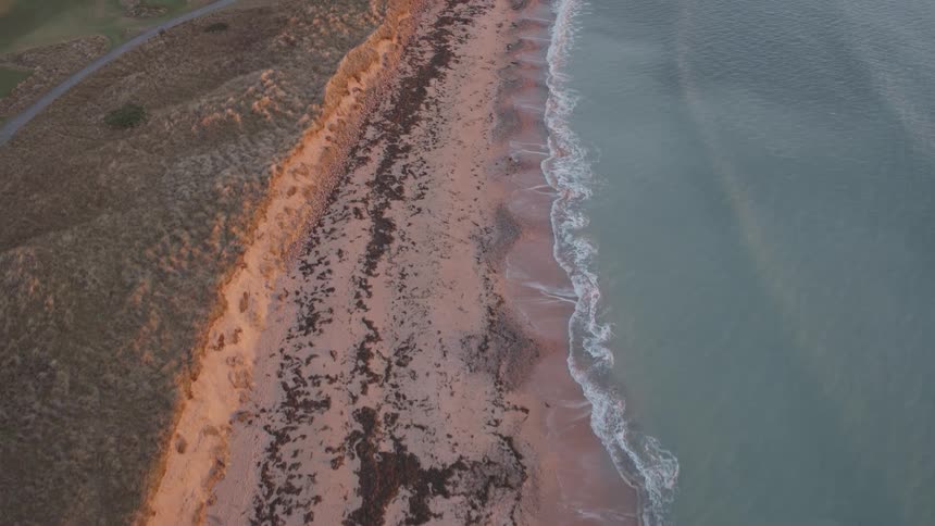 An aerial view follows the serene coastline where gentle waves meet a sandy beach next to golden-lit dunes at sunrise