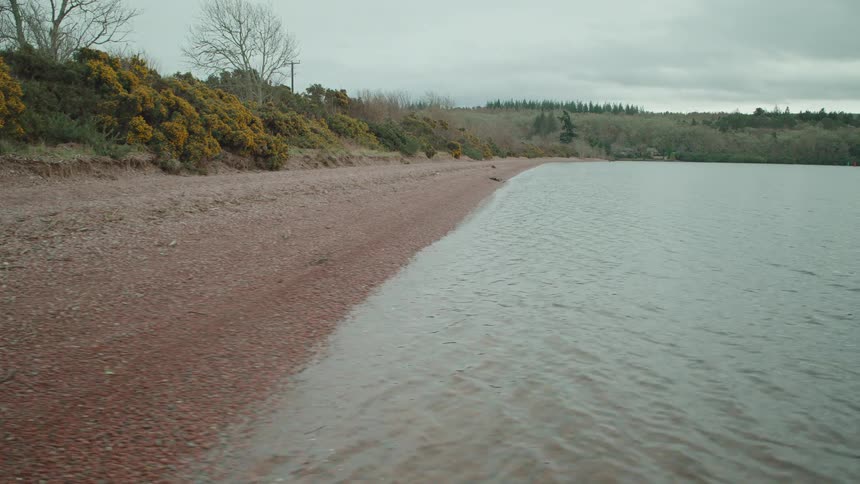 A serene low-angle tracking shot pulling back from a pebble beach over the tranquil water of a Scottish loch on a cloudy day