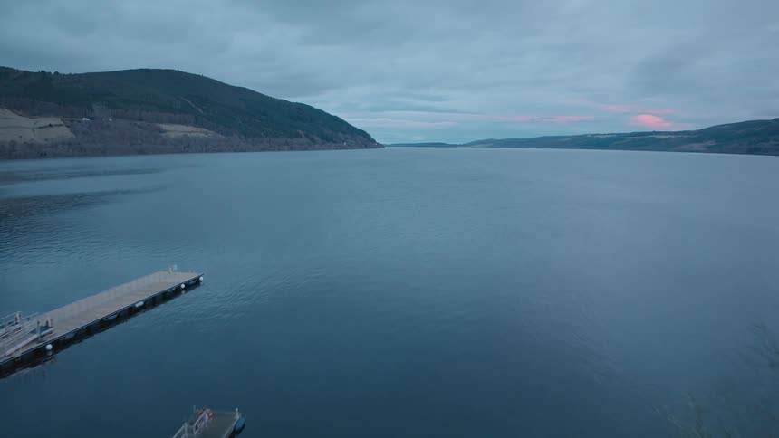 Aerial view flying over a grassy park towards the calm waters of a vast loch at dusk