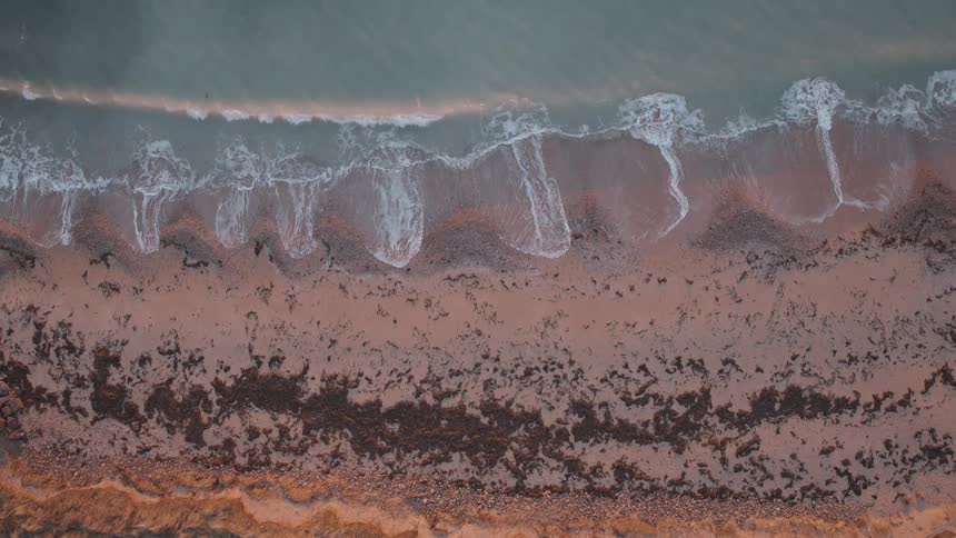 A serene top-down aerial view of ocean waves washing over a pink sandy shore in slow motion