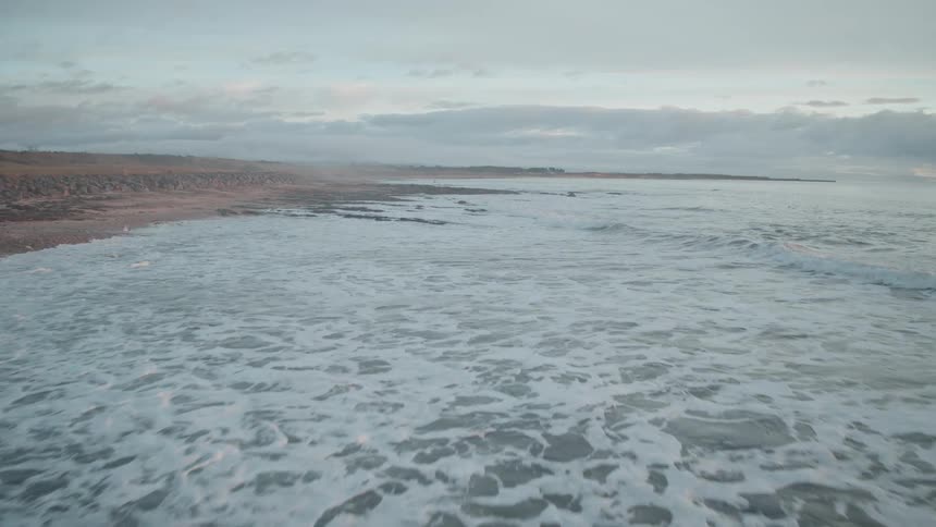 A slow aerial shot moves from a rocky beach out over the foamy ocean waves at dusk