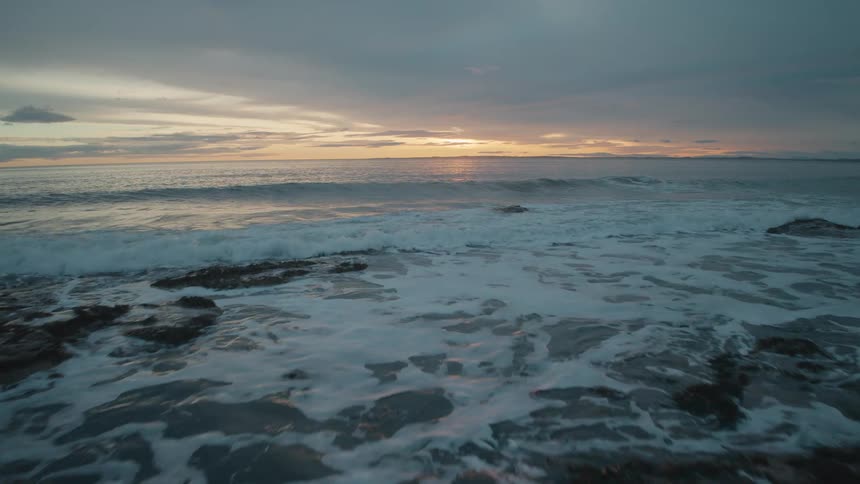 A slow, cinematic dolly shot moves from a rocky beach over the incoming tide and out to the open ocean at sunset