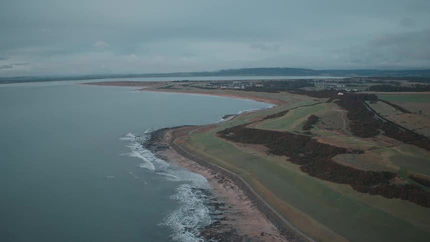 An aerial view slowly ascends over a tranquil coastline, revealing a lush green landscape and distant town under an overcast sky